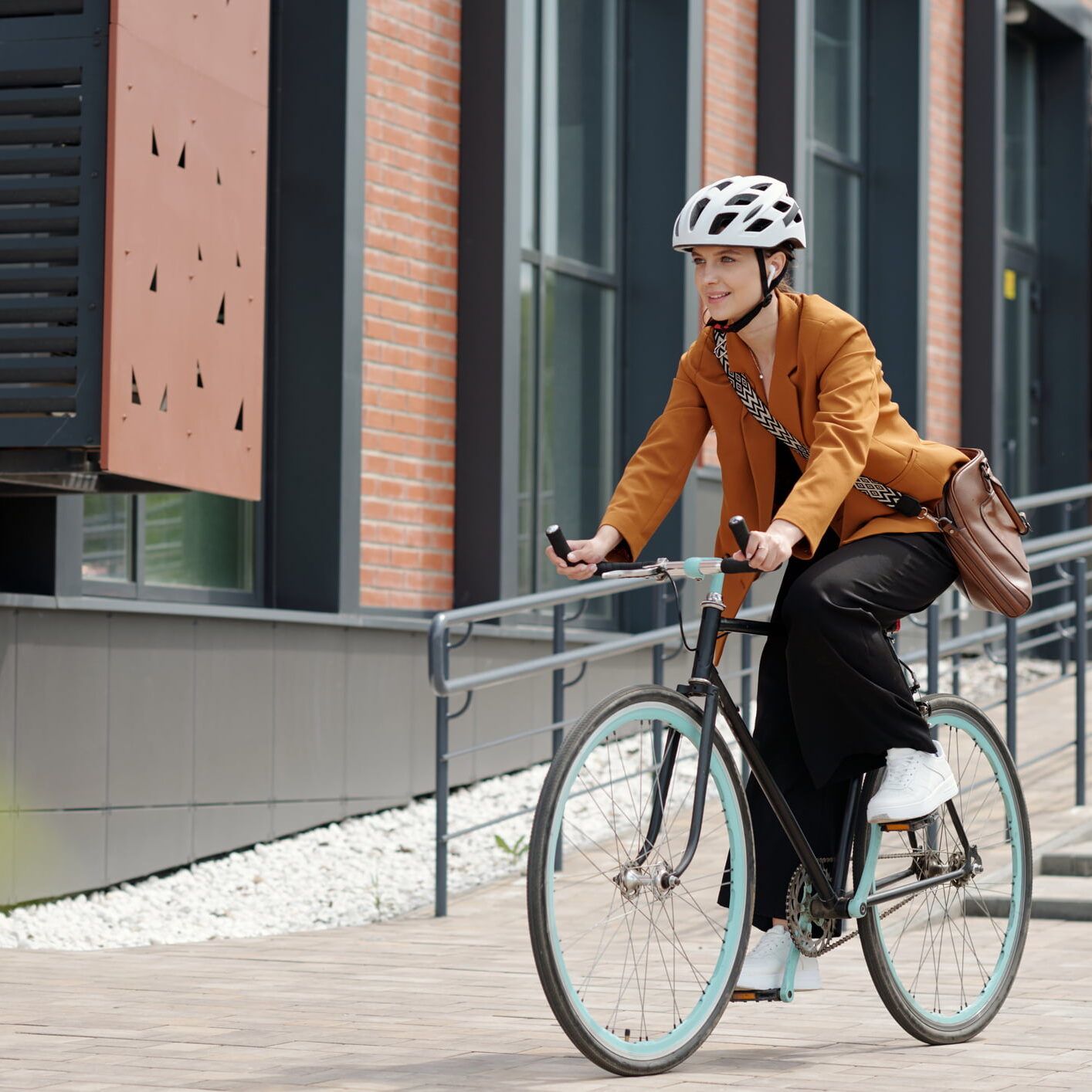 Young woman in formalwear and safety helmet sitting on bicycle and riding home from work while moving along modern building